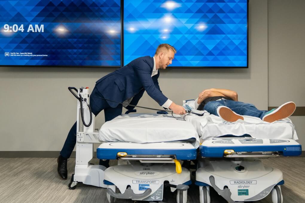 Man demonstrates SimPull device. Two beds are next to each other, one holds a person pretending to be a patient. The demonstrator is putting straps under the patient. The SimPull device will help move a patient from one bed to another.